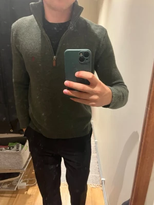 Mörkgrön halvzip tröja - Mörkgrön stickad tröja med halvzip och hög krage. Tröjan har ribbade muddar vid ärmslut och nederkant samt en liten broderad logga på bröstet. Perfekt för lager-på-lager och snygg till både jeans och chinos.