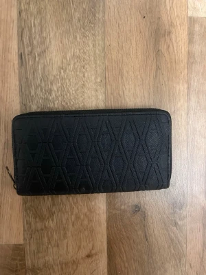 Svart plånbok med mönster - Snygg svart plånbok med dragkedja runtom och geometriskt mönster på utsidan. Plånboken har flera fack för kort och ett myntfack med dragkedja inuti. Tillverkad i textilmaterial med stilren design.