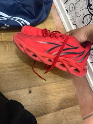 Röda sneakers med unik sula - Säljer ett par riktigt snygga röda sneakers med futuristisk, hålig sula och svarta detaljer på sidan. Skorna har snörning och är tillverkade i mesh och syntetmaterial för en sportig vibe. Perfekta för dig som vill sticka ut med din stil.