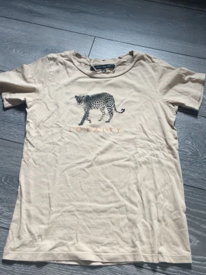 Beige t-shirt med leopard från KappAhl - Snygg beige t-shirt från KappAhl i mjuk bomull. Framtill finns ett leopardtryck och texten 'LOYALTY' i orange. Klassisk rund halsringning och korta ärmar. Perfekt för dig som gillar djurmotiv och enkel stil.