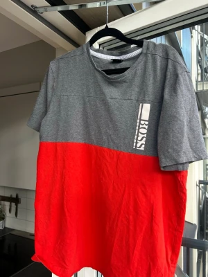 Grå och röd t-shirt från Hugo Boss - Snygg t-shirt från Hugo Boss med grå överdel och röd nederdel. T-shirten har rund halsringning, korta ärmar och en vertikal logga på bröstet. Tillverkad i mjuk bomull för skön känsla och stilren look.