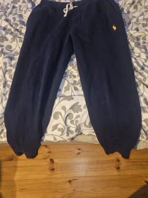 Mörkblå mjukisbyxor Polo Ralph Lauren - Mörkblå mjukisbyxor från Polo Ralph Lauren med dragsko i midjan och gul broderad logga på vänster sida. Byxorna har muddar vid bensluten och är gjorda i mjukt, fleeceliknande material. Perfekta för chill dagar eller när du vill vara extra bekväm.