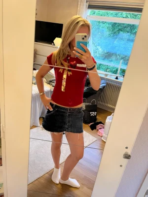 Röd topp med paljettrosett - Snygg röd topp med korta ärmar och en stor guldfärgad paljettrosett framtill. T-shirten har en tight passform och är perfekt för dig som vill sticka ut lite extra. Materialet känns mjukt och stretchigt, troligen bomull eller polyester.
