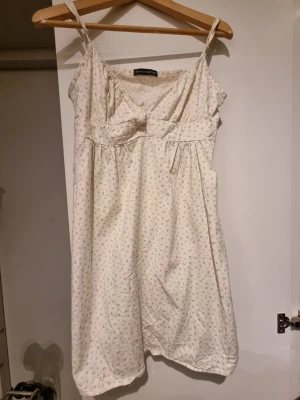 Vit blommig kortklänning med knyt från Brandy Melville - Söt vit kortklänning med tunna axelband och små blommor i mönstret. Klänningen har en feminin passform, knyt i ryggen och lätt v-ringad hals. Perfekt för varma dagar och sommarens alla tillfällen.