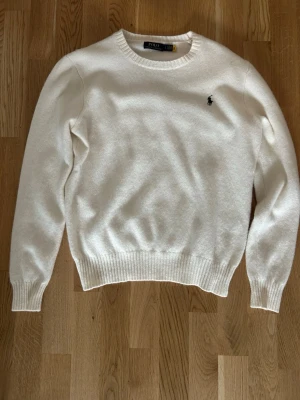 Vit stickad tröja från Polo Ralph Lauren - Klassisk vit stickad tröja från Polo Ralph Lauren med rund halsringning och broderad logga på bröstet. Tröjan har ribbstickade muddar vid ärmslut och nederkant. Perfekt för en stilren look.