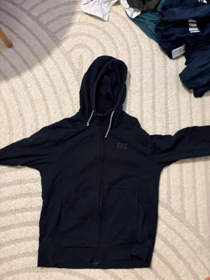 Svart zip hoodie från EA7 - Snygg svart hoodie från EA7 med dragkedja framtill och två fickor. Har huva med dragsko och diskret EA7-logga på bröstet. Perfekt för en chill och sportig look. Materialet känns mjukt och bekvämt.