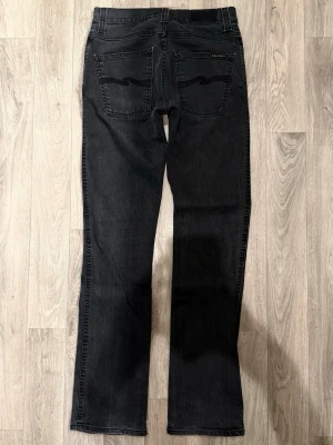 Nudie Jeans Slim Jim Mörkgrå W31/L34 - Slim Fit - Nudie Jeans Slim Jim ”Dry Black Coated” mörkgrå jeans med slim fit-passform, normal midja och lätt bootcut, storlek W31/L34, midja 37,5 cm, ytterbenslängd 107,5 cm, benöppning 19 cm, modellen är 185cm & väger 65kg, i fint skick.