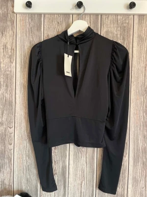 Trendig svart långärmad topp med puffärm S helt ny - Trendig svart topp med lång puffärm och hög hals. Toppen har en snygg öppning framtill och rynkade detaljer vid midjan som ger en cool siluett. Perfekt för dig som gillar stilrena plagg med en twist.