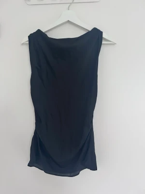 Svart topp från Zara - Säljer en stilren svart topp från Zara. Toppen är ärmlös med en mjuk, draperad halsringning och har en kort längd. Materialet känns lätt och har en sidenliknande finish som ger en elegant look.