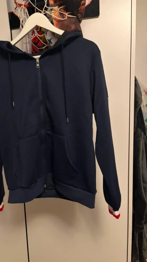  Moncler hoodie  - Snygg marinblå Moncler  hoodie med dragkedja framtill och huva med dragsko. Fickor på magen och ribbade muddar med vita och röda detaljer vid ärmsluten. Perfekt för en chill och sportig look.