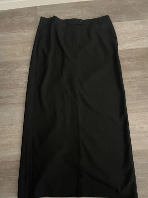 Svart långkjol med slits - Stilren svart randig kostymkjol med diskreta bälteshällor och slits baktill. Kjolen har en rak passform och stängs med knapp och dragkedja i midjan. Perfekt för en clean och klassisk look.