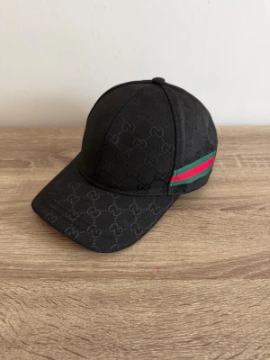 Keps - Svart keps med snyggt monogram över hela tyget och klassiska gröna och röda ränder på sidorna. Justerbar rem baktill i läder. Snygg och stilren accessoar i canvasmaterial, perfekt för att lyfta din outfit. Helt ny oanvänd. 