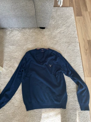 Mörkblå stickad tröja från GANT - Mörkblå stickad tröja från GANT med V-ringning och broderad logga på bröstet. Tillverkad i mjuk merinoull och har långa ärmar samt ribbade muddar vid ärmslut och nederkant. Perfekt för en stilren och klassisk look.