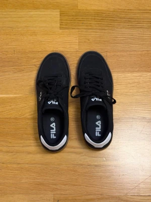Svarta Fila sneakers med gummisula - Svarta sneakers från Fila med klassisk låg profil och vit detalj på hälen. Skorna har svart ovandel i textil, svart snörning och en brun gummisula. Fila-logga syns på sidan och plösen. Perfekta för en sportig och avslappnad stil.
