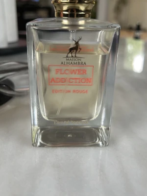 Flower Addiction Edition Rouge parfym - Maison Alhambra Flower Addiction Edition Rouge Eau de Parfum i en elegant, genomskinlig glasflaska med guldlock. Doften har en modern och fräsch känsla, och flaskan har en stilren, fyrkantig form med röd text på framsidan. 75 ml.