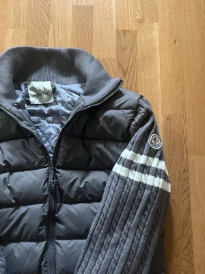 Grå dunväst från Moncler - Snygg grå dunväst från Moncler med stickade ärmar och ribbad krage. Västen har dragkedja framtill och Moncler-logga på ärmen. Ärmarna har vita ränder och är ribbstickade för extra stilpoäng. Perfekt för dig som vill ha en sportig och trendig look. Jackan är i Storlek M. Jackan är stor i armarna på mig men generella längden är bra! Skriv för fler frågor/ funderingar!