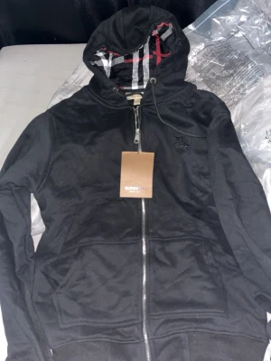 Burberry Kofta - Snygg svart hoodie från Burberry med dragkedja framtill och klassiskt rutigt foder i huvan. Hoodien har fickor på magen och justerbar dragsko i huvan. Perfekt för dig som vill ha en stilren och exklusiv look. Materialet känns mjukt och bekvämt.