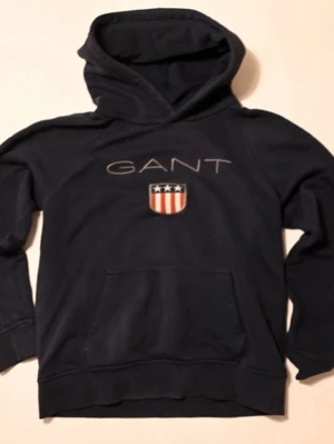 Mörkblå hoodie från GANT 152 - Snygg mörkblå hoodie från GANT med stor huva och känguruficka. Framtill finns GANT-logga och ett sköldmärke med stjärnor och ränder i rött, vitt och blått. Perfekt för en avslappnad och stilren look.