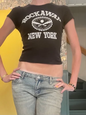 Svart croppad t-shirt Rockaway - Säljer en svart croppad t-shirt med trycket 'Rockaway New York' i vitt framtill. T-shirten har korta ärmar och en tight passform. Perfekt för dig som gillar streetstyle och vill ha en cool statement-tee i garderoben.