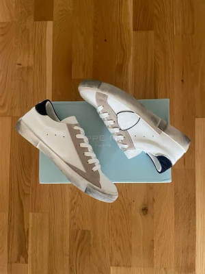 Vita och beige sneakers från Philippe Model - Snygga sneakers från Philippe Model i vitt läder med beige mockadetaljer och svart hälparti. Klassisk låg modell med vita snören och slitstark gummisula. Perfekta för en clean och stilren look till vardagsoutfiten.
