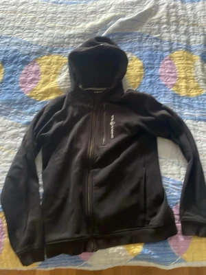 Svart hoodie med dragkedja - Säljer en svart hoodie med dragkedja och huva. Hoodien har en bröstficka med dragkedja och diskret tryck på bröstet. Perfekt för chill dagar eller när du vill ha en enkel och stilren look. Materialet känns mjukt och bekvämt.