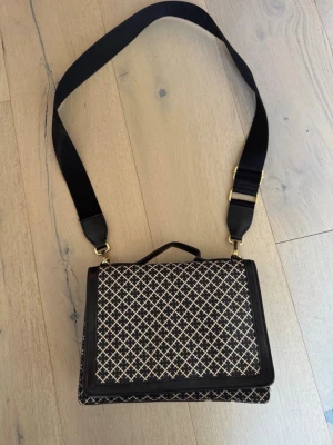 Malene Birger väska - Säljer en Malene Birger väska Snygg axelväska i svart och beige med rutigt mönster och bred axelrem. Väskan har en fyrkantig form, lockstängning och detaljer i svart konstläder. Perfekt för dig som vill ha en unik accessoar med cool design. 