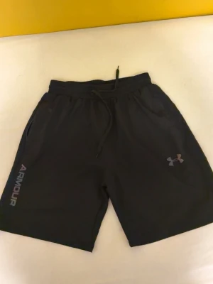 Svarta träningsshorts från Under Armour - Svarta shorts från Under Armour med snörning i midjan och logga på vänster ben. Passar som S Tillverkade i ett lätt och bekvämt material som passar perfekt för träning. Klassisk passform med två sidofickor och elastisk midja.