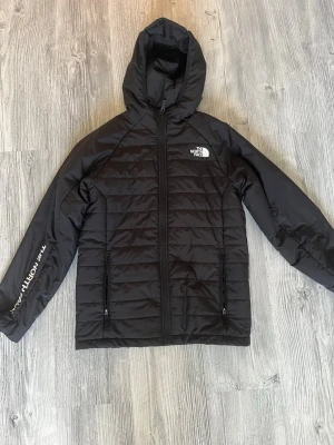 Svart vindjacka från The North Face - Snygg svart vindjacka från The North Face med huva och dragkedja framtill. Jackan har två fickor med dragkedja och logga på bröstet samt ärmen. Tillverkad i lätt och slitstarkt material, perfekt för blåsiga dagar.