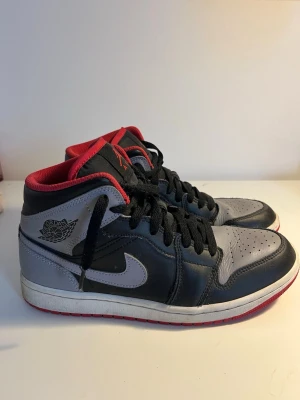 Air Jordan svart/grå/röd sneakers - Snygga Air Jordan sneakers i svart och grått med röda detaljer och svart snörning. Skorna har klassisk Jordan-logga på sidan och tungan, samt perforerad tå för extra ventilation. Yttermaterial i läder och textil, med vit sula och röd undersida, skorna är använda ungefär två gånger