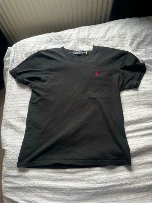 Svart t-shirt från Ralph Lauren - Klassisk svart t-shirt från Ralph Lauren med korta ärmar och rund hals. Liten röd broderad logga på bröstet ger en stilren touch. Tillverkad i mjuk bomull för skön känsla och enkel matchning till olika outfits.