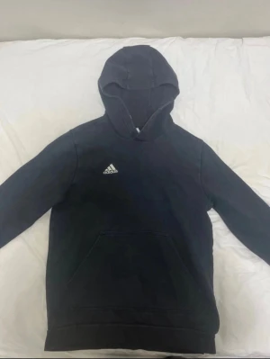 Svart hoodie från Adidas med huva - Snygg svart hoodie från Adidas med klassisk huva och stor magficka framtill. Adidas-loggan sitter på bröstet. Hoodien har långa ärmar och ribbade muddar vid ärmslut och nederkant. Perfekt för en avslappnad och sportig stil.