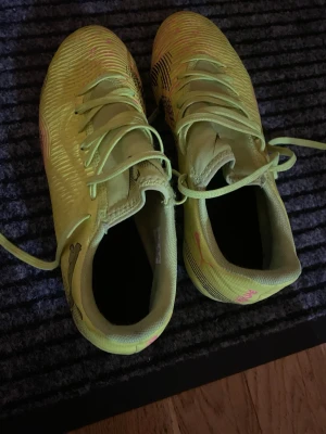 Gula Puma fotbollsskor med dobbar - Säljer ett par gula fotbollsskor från Puma med svarta och rosa detaljer. Skorna har snörning och dobbar i plast, perfekta för spel på gräsplan. Snygg design med mönstrade detaljer på sidorna och klassisk Puma-logga.