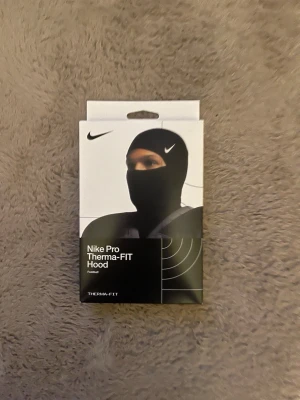 Nike Pro Therma-FIT balaclava - Nike Pro Therma-FIT balaclava i svart med vit Nike-logga. Tillverkad i ett mjukt och stretchigt material som håller värmen och andas. Perfekt för kalla dagar och sportaktiviteter utomhus. Snygg och funktionell design som täcker hela huvudet och halsen.