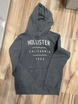 Grå hoodie från Hollister - Mysig grå hoodie från Hollister med tryck på bröstet där det står 'Hollister California 1922'. Klassisk känguruficka framtill och justerbar huva. Perfekt för chill dagar och enkel att matcha med jeans eller mjukisbyxor.