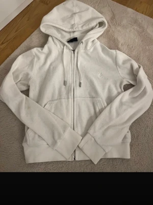 Vit hoodie från Juicy Couture - Säljer en vit hoodie från Juicy Couture med dragkedja framtill, huva med dragsko och fickor på sidorna. Klassisk och enkel design med diskret logga på bröstet. Mjuk och skön bomullsblandning som passar perfekt till chill dagar.