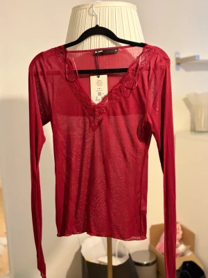 Vinröd mesh topp med spetsdetaljer - Snygg vinröd mesh topp med långa ärmar och v-ringning. Spetsdetaljer vid halsringning och ärmslut ger en extra touch. Tunn och genomskinlig, perfekt att styla lager på lager. Materialet är lätt och stretchigt.