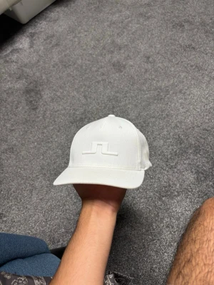 Vit J. Lindeberg Keps - Snygg oanvänd vit J. lindeberg keps med böjd skärm och broderad logotyp framtill. Justerbar snapback baktill för perfekt passform. Kepsen har en stilren och minimalistisk design som passar till många outfits.