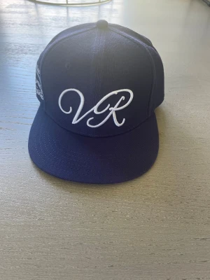 Mörkblå snapback keps med VR-brodyr - Snygg mörkblå snapback keps med vit brodyr framtill där det står 'VR' i stilren skrivstil. Klassisk platt skärm. Perfekt accessoar för en streetwear-look. Materialet ser ut att vara bomull eller polyester. Priset kan diskuteras vid snabb affär 🔜storleken är cirka 55cm