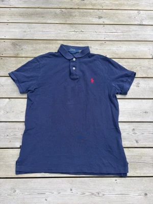 Marinblå pikétröja från Ralph Lauren - Klassisk marinblå pikétröja från Ralph Lauren med röd broderad logga på bröstet. Tröjan har korta ärmar, krage och två knappar framtill. Slim fit-modell i bomull som passar perfekt till jeans eller shorts.