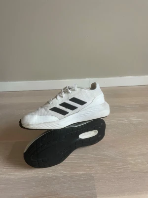Vita Adidas träningsskor med svarta ränder - Snygga vita sneakers från Adidas med klassiska svarta ränder på sidan. Skorna har en mesh-liknande ovandel och tjock vit sula med svart yttersula. Perfekta för en sportig och avslappnad stil.