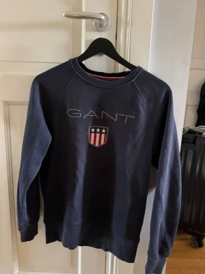 Mörkblå sweatshirt från GANT - Mörkblå sweatshirt från GANT med broderad logga och amerikanskinspirerat sköldmärke på bröstet. Tröjan har rund halsringning, långa ärmar och ribbade muddar vid ärmslut och nederkant. Perfekt för en avslappnad och stilren look.