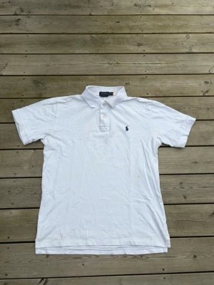 Vit piké från Polo Ralph Lauren - Klassisk vit pikétröja från Polo Ralph Lauren med korta ärmar och blå broderad logga på bröstet. Tröjan har krage och två knappar framtill. Tillverkad i bomull för en skön och luftig känsla.