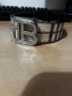 Rutmönstrat beige bälte med B-spänne - Snyggt bälte med klassiskt rutmönster i beige, svart och rött. Bältet har ett stort silvrigt B-format spänne som ger en lyxig känsla. Perfekt accessoar för att lyfta din outfit och ge den en trendig touch.