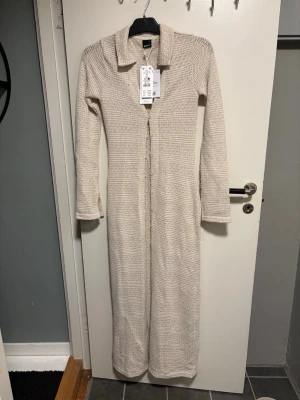 Beige virkad maxiklänning Gina Tricot - Lång beige maxiklänning från Gina Tricot i virkad stil. Klänningen har lång ärm, krage och knäppning framtill. Tillverkad i ett mjukt, återvunnet material som ger en avslappnad men trendig look.