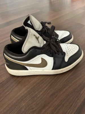 Air Jordan 1 Low - Snygga Nike Air Jordan 1 Low sneakers i svart och vitt med bronsfärgad swoosh. Skorna har klassisk låg siluett, perforerad tåbox och svarta skosnören. Tillverkade i läder med textildetaljer och ikoniska Jordan-loggor på plös och häl. Köpta för 1300kr