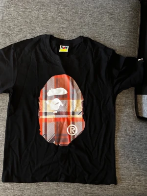 Svart t-shirt med rutig Bape-logga - Svart t-shirt med stor ikonisk Bape-logga framtill i röd, beige och svart rutig design. Klassisk passform och rund halsringning. T-shirten har en liten etikett med logga på ärmen. Perfekt för dig som gillar streetwear.