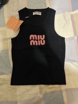 Svart ärmlös linnetopp från Miu Miu - Snygg svart linnetopp från Miu Miu med pärlbroderad logga i vitt och rött framtill. Toppen är ärmlös och har en klassisk rund halsringning. Perfekt för dig som vill ha en trendig och exklusiv look.