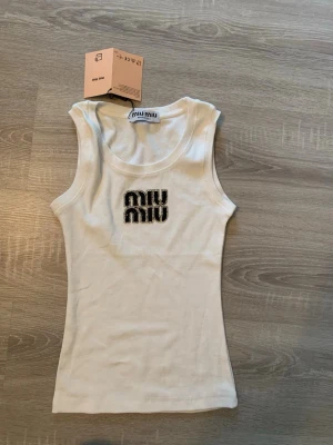 Vit ribbad linnetopp från Miu Miu - Säljer en vit ribbad linnetopp från Miu Miu med logga framtill i svart och silverdetaljer. Toppen är ärmlös och har en tight passform, perfekt för en trendig och enkel look.