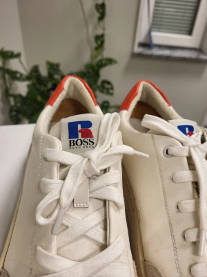 BOSS x Russell Athletic Amber low-top sneakers in white and cream leather 42 - Snygga vita sneakers från Hugo Boss med klassisk låg profil. Skorna har vita snören, beige mockadetaljer vid tån och en röd detalj på hälen. Märkets logga syns tydligt på plösen. Ovandelen är i läder och mocka för en clean look.