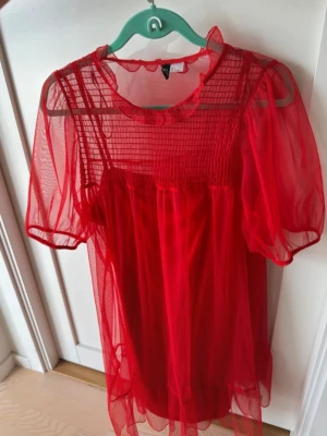 Röd meshklänning från H&M Divided - Säljer en röd meshklänning från H&M Divided med volangkant vid halsen och transparenta korta ärmar. Klänningen har ett rutigt mönster upptill och är gjord i ett lätt, syntetiskt material. Perfekt för dig som vill sticka ut med en unik look.